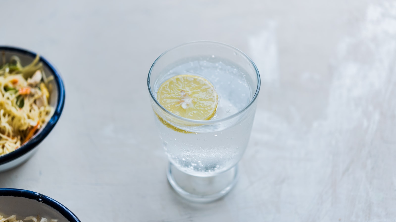 Sklenice čisté vody s plátkem citronu vedle misky jídla na světlém stole